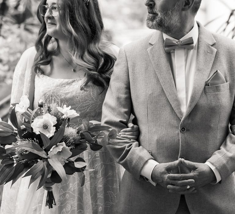 Father of the bride in a grey wedding suit stands linking arms with the bride as they prepare to walk down the aisle together 