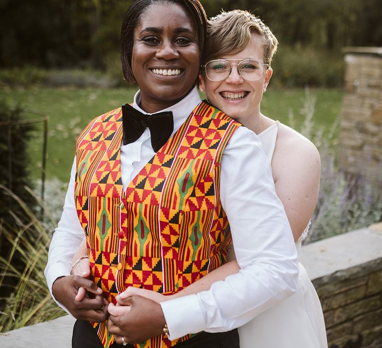 Bride in bold Ghanian patterned waistcoat being embraced from behind by bride in a white wedding dress 