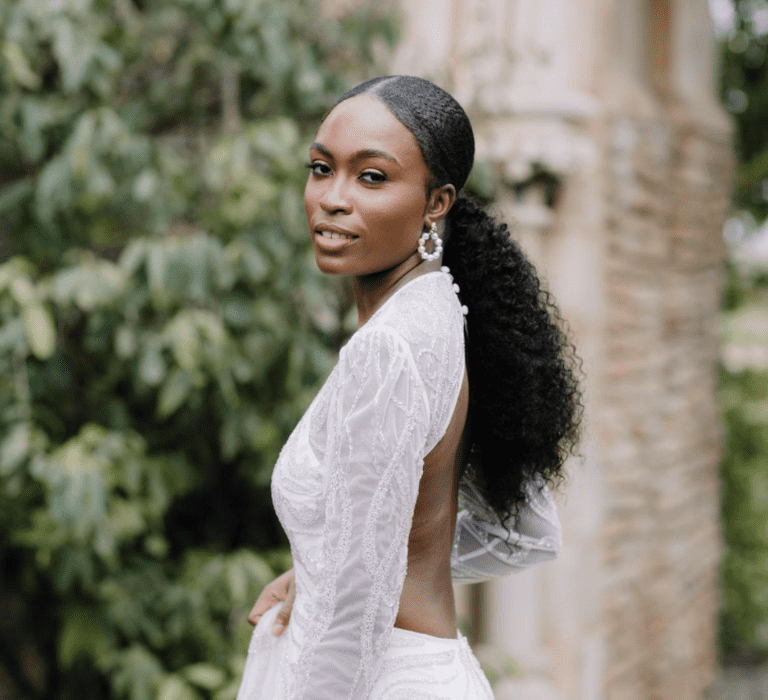 A Black bride wears a long sleeved backless wedding dress. She has her hair in a low ponytail.