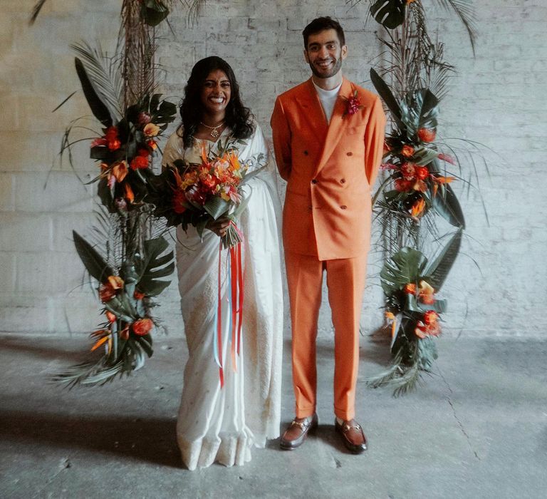 Asian bride in a white sari and Turkish groom in an orange double breasted suit standing at the altar decorated in tropical wedding flowers 