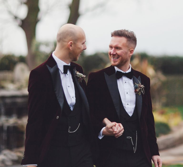 LGBTQ+ couple holding hands in wine coloured velvet tuxedo jackets 