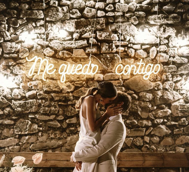 Groom in a beige suit picking up his bride in a slip dress in front of a neon sign 