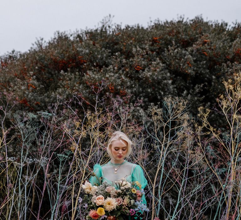 Stylish bride in a sheer tulle pastel wedding dress standing in front of a colourful flower wall holding a bright wedding bouquet tied with ribbon