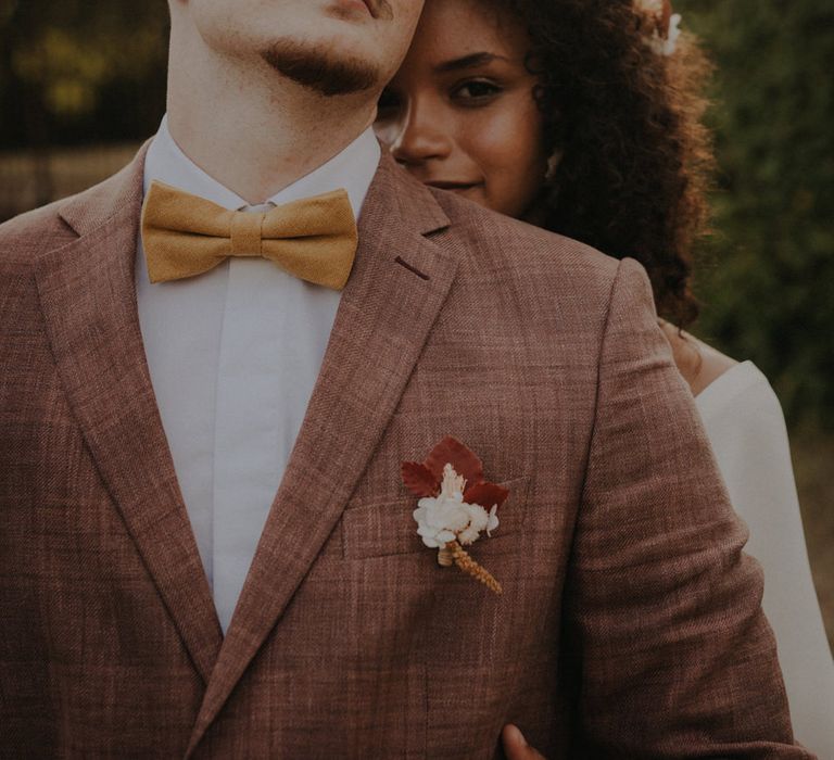 Bride with naturally curly hair peeking over her groom's shoulder in a brown check suit and ochre bow tie