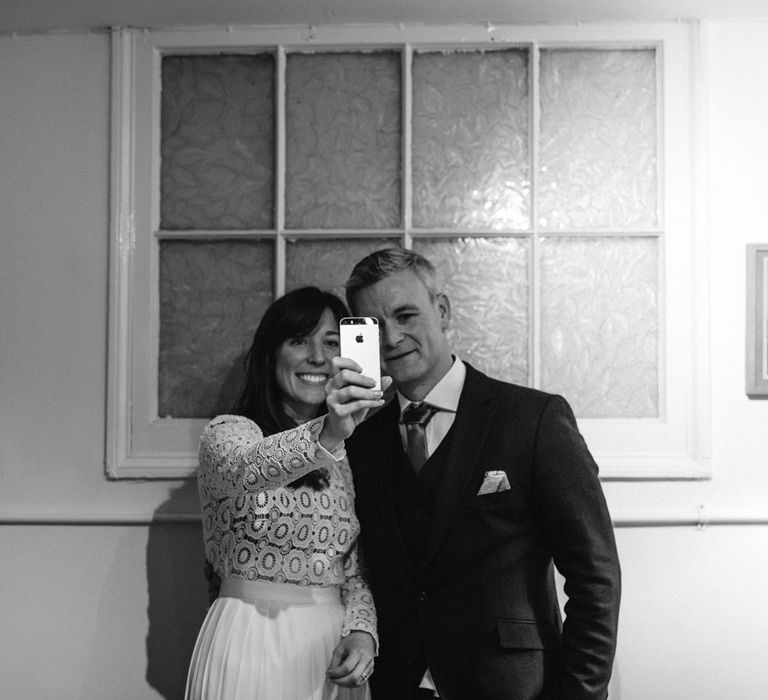 Bride in long sleeved lace top Self Portrait wedding dress stands next to groom in brown woollen blazer holding up phone whilst FaceTiming family and friends 