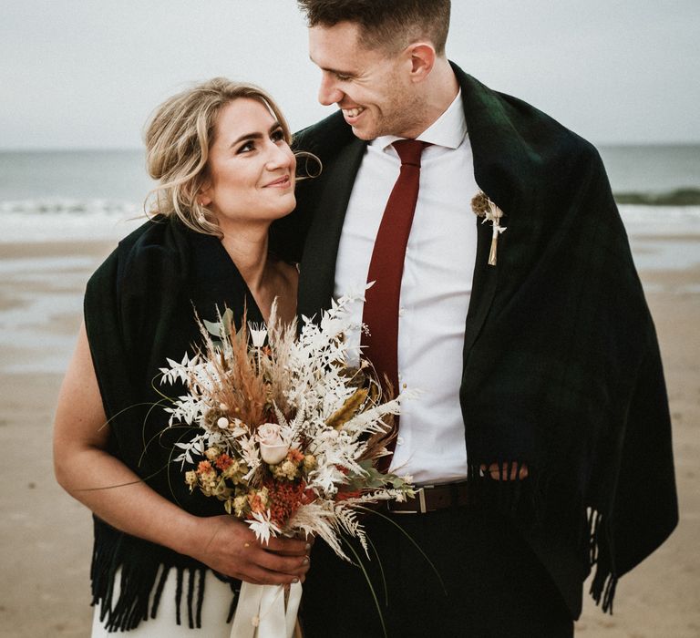 Bride & groom cover themselves in shawl on the beach as they look at one another lovingly 