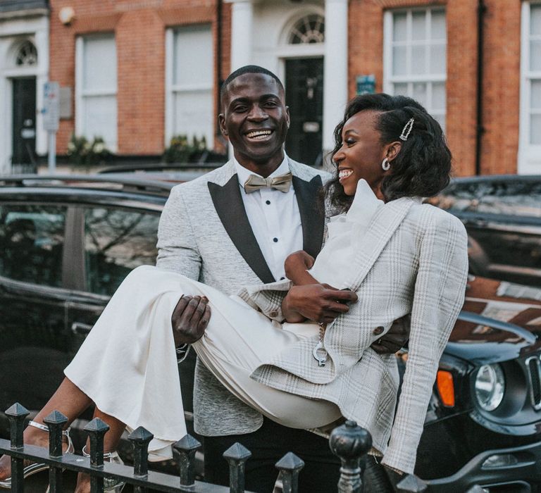 Laughing groom in grey suit jacket with black lapels picking up smiling bride in white satin dress and grey checkered blazer on a street in Leeds