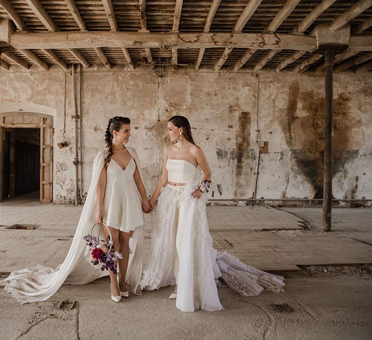 Two brides holding hands in an industrial venue in a mini wedding dress holding a hoop bouquet and bridal separates with detachable ruffle skirt 