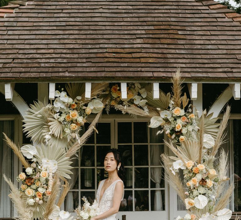 Bride at Berwick lodge wearing a plunge neck wedding gown with tulle skirt, under an arch of dried palms, pampas grass, white anthuriums, roses and orchids