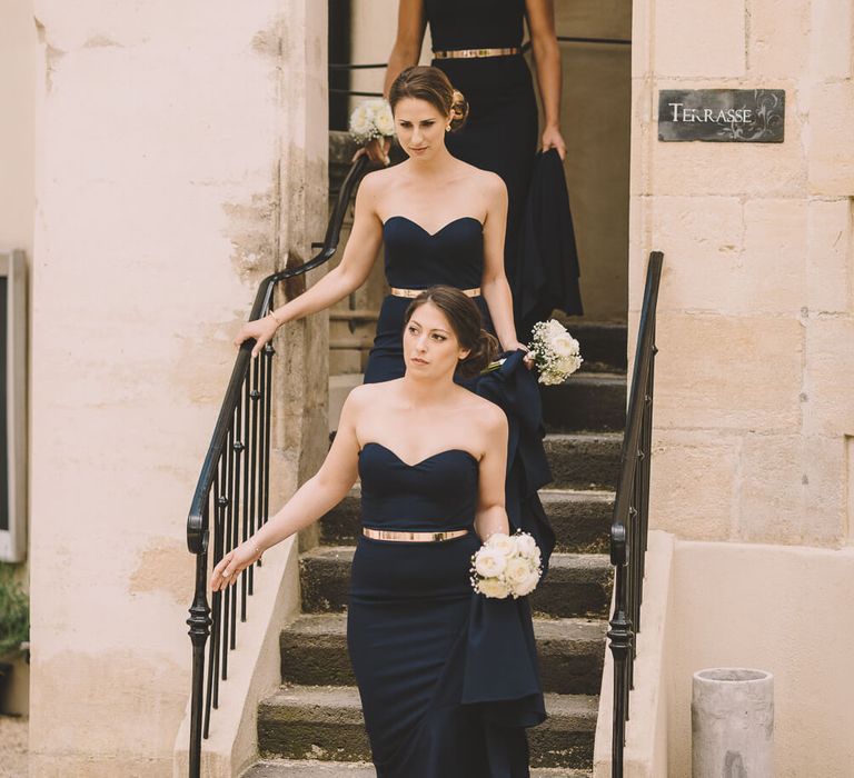 Bridesmaids walking down a staircase wearing strapless navy bridesmaid dresses with gold belt | White Runway Dresses | Photography by Modern Vintage Weddings