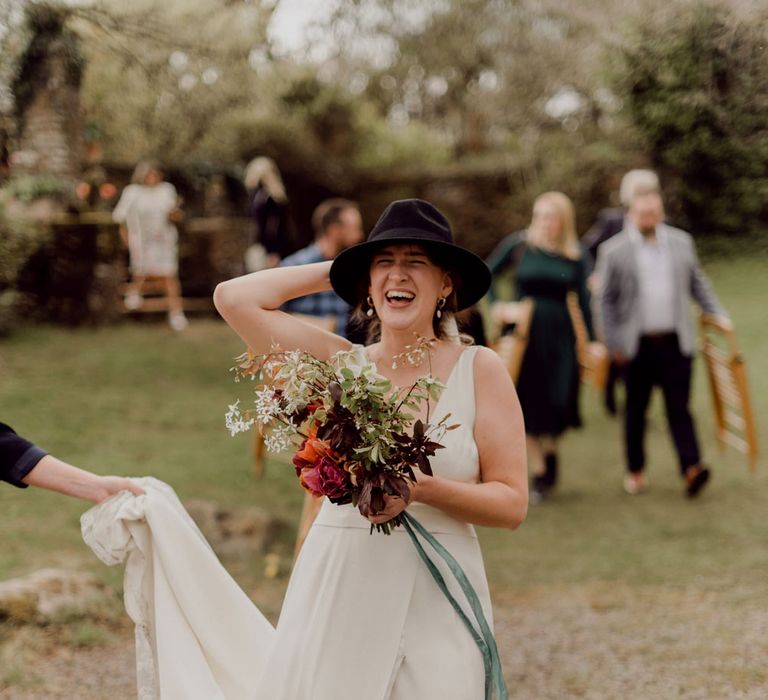 Laughing bride in Charlie Brear wedding dress and black fedora walks whilst holding multicoloured wedding bouquet at garden party wedding in Devon