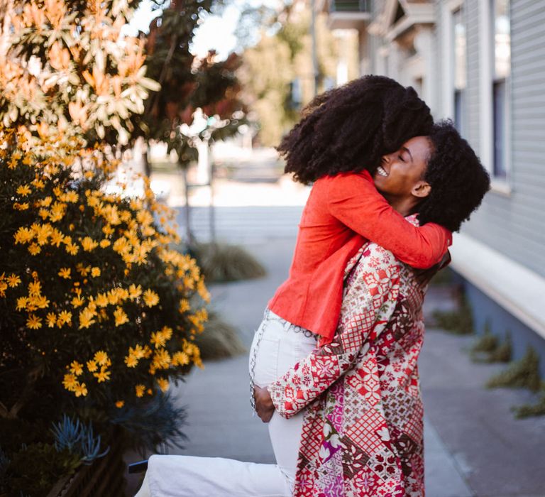 Bride-to-be in a long line patterned jacket embracing her bride-to-be in white jeans and a burnt orange jacket 