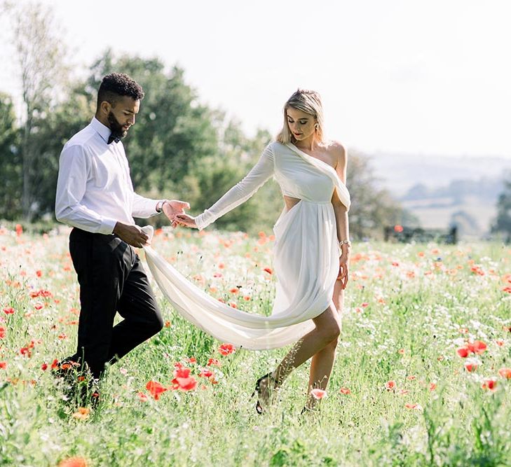 Groom in a white shirt and bow tie holding his brides wedding dress train as they walk through poppy fields 