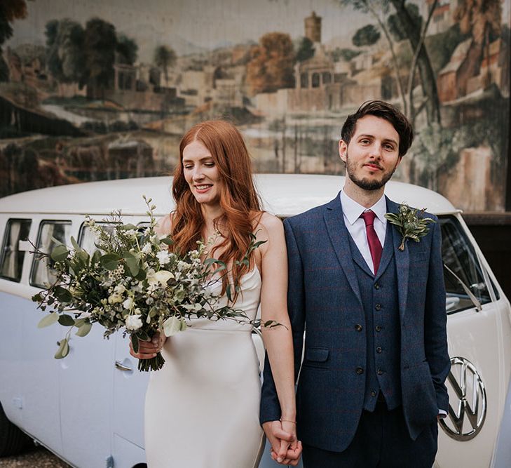 Groom in a three-piece navy blue wool suit holding hands with his bride in a satin slip wedding dress standing in front of a pastel blue VW camper van 