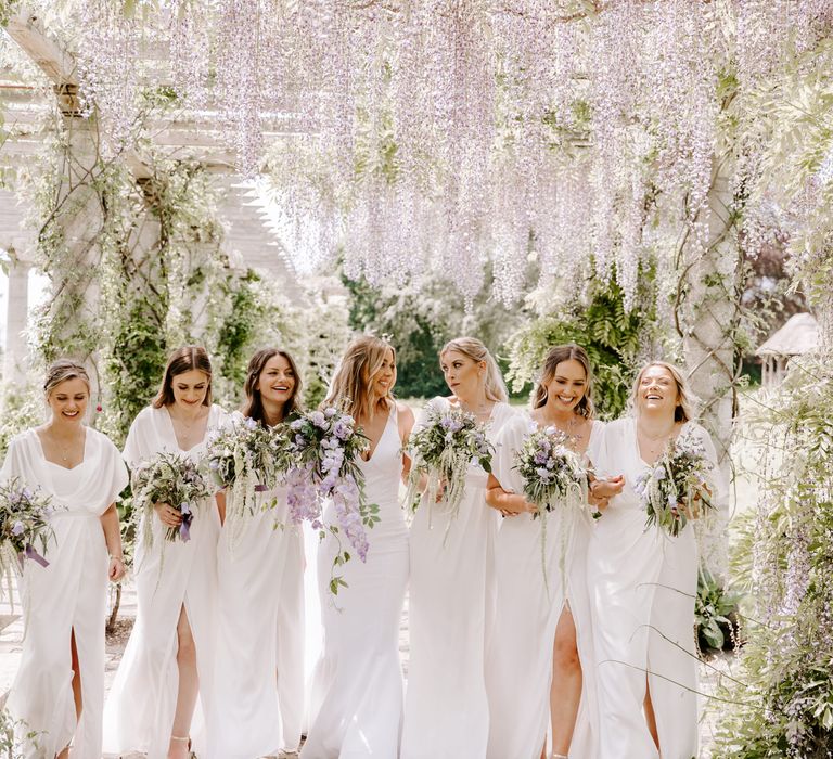 Bride stands with her bridesmaids who wear white ASOS dresses beneath pergola with lilac hanging florals