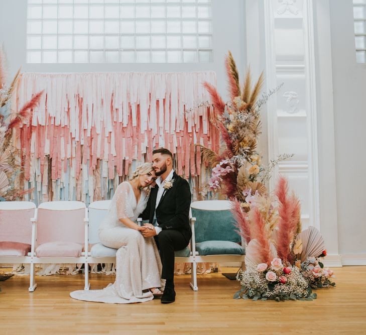 Bride and groom sitting on a row of velvet pastel wedding chairs in front of a tissue paper backdrop flanked by vertical flower arrangements 