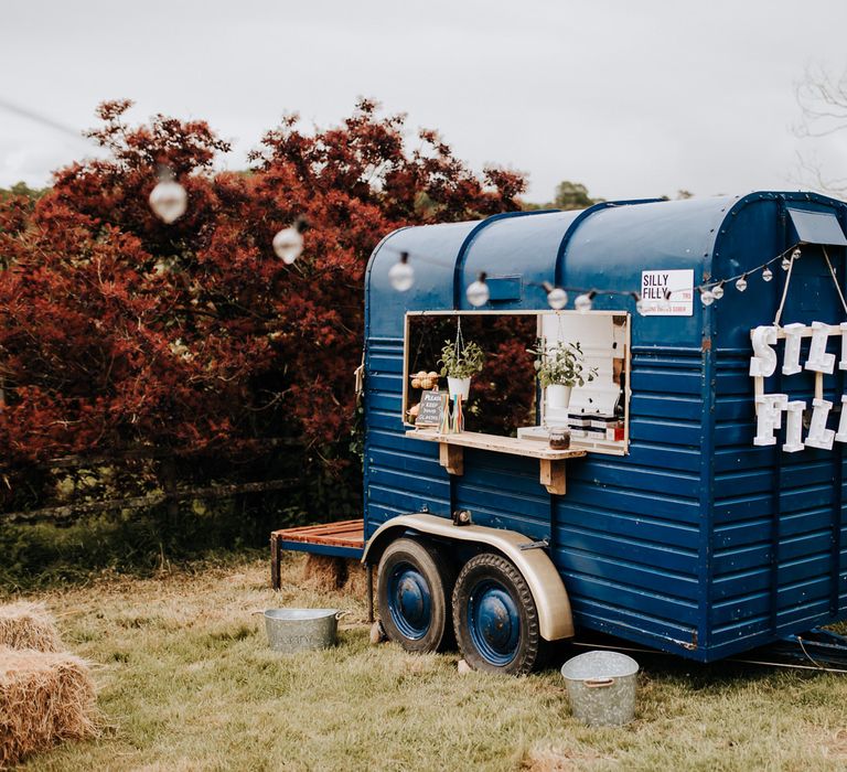 Dark blue horse box bar in field with lightbulb fairy lights, 'Silly Filly' sign and hanging plants for garden wedding reception