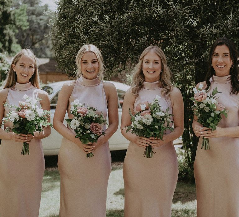 Four bridesmaids stand outside in blush pink high necked satin bridesmaids dresses holding green, white and pink wedding bouquets before Surrey summer wedding