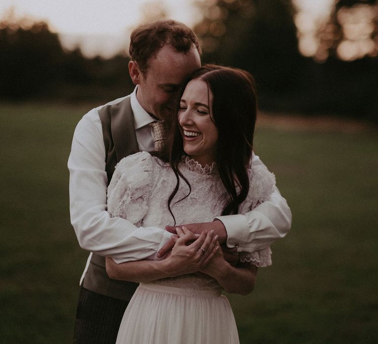 Groom in white shirt and green waistcoat hugs bride in white lace puffed sleeve Daarlana wedding dress from behind as she laughs in the grounds of Loseley Park during golden hour