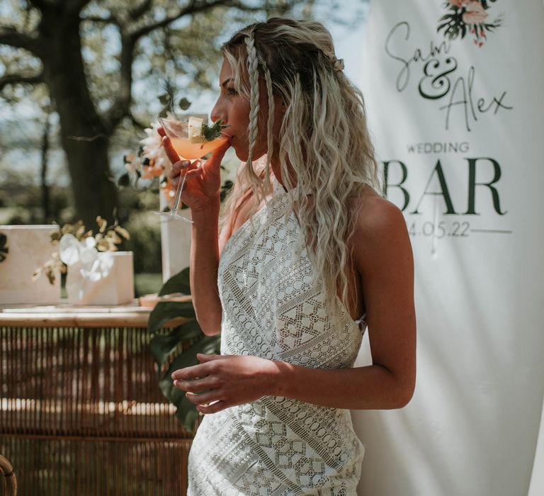 Bride with blonde beach waves and plaits wearing white lace boho wedding dress sips orange cocktail from a martini glass in front of surfboard bar sign for birds of paradise wedding inspiration