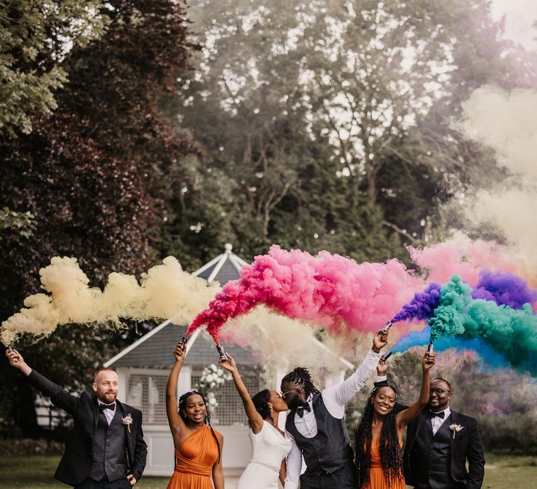 Smoke bomb portrait of the bride and groom kissing as the bridesmaids in orange dresses and groomsmen hold the coloured grenades 