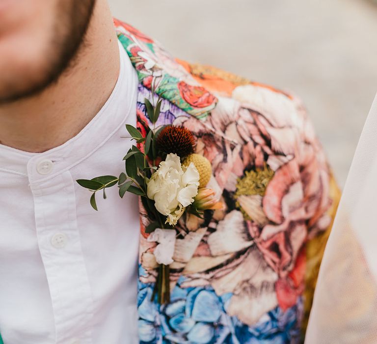 Groom in a white grandad collar shirt, colourful patterned blazer and white, red and yellow button hole flowers 