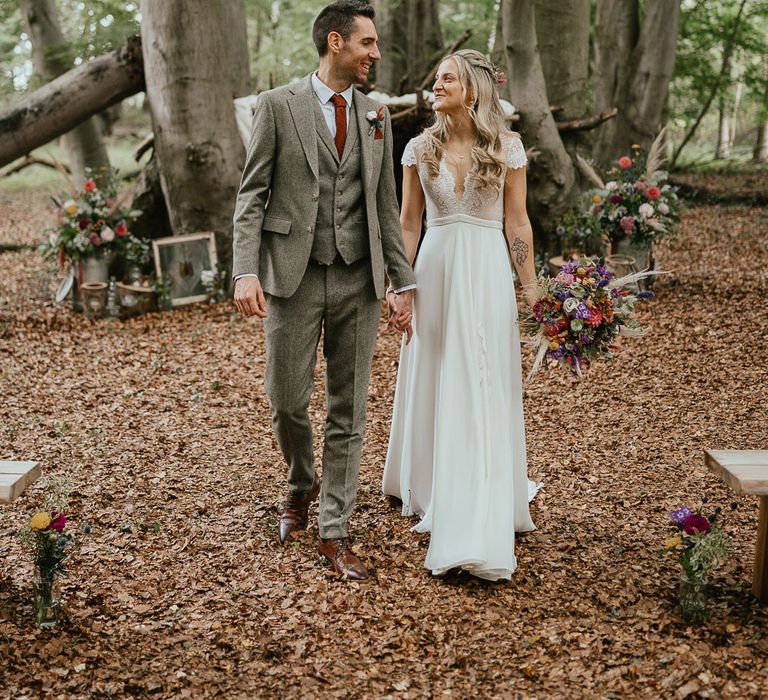 Groom in grey herringbone suit and red tie laughs with bride in white lace cap sleeved wedding dress holding mixed bridal bouquet in woodland at late summer wedding in Norfolk