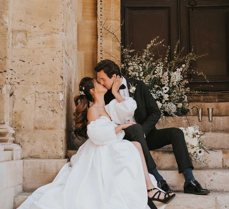 Bride in a stylish wedding dress with front split and detachable puff sleeves kissing her groom on the steps at Bodleian wedding venue in Oxford