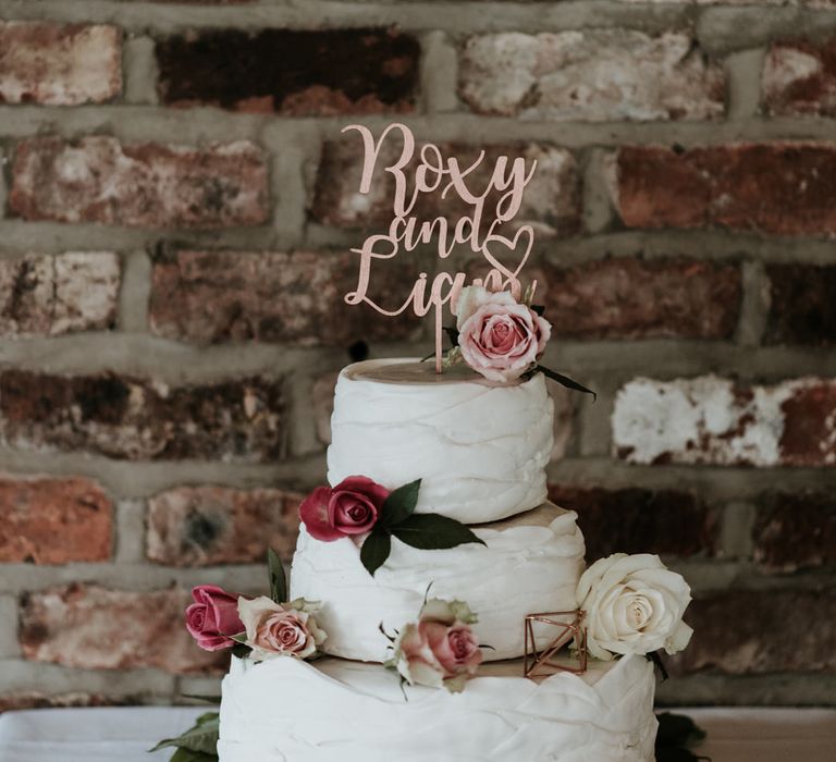 three-tier buttercream wedding cake with floral decor and rose gold wedding cake topper of the bride and grooms names 