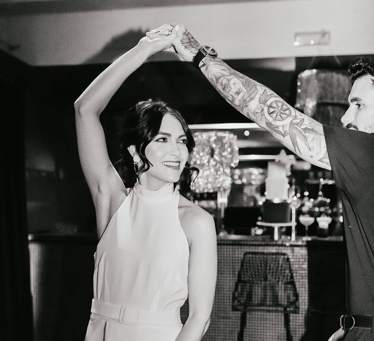 Black and white portrait of the groom in a black short sleeve shirt twirling his bride in a halterneck wedding dress 