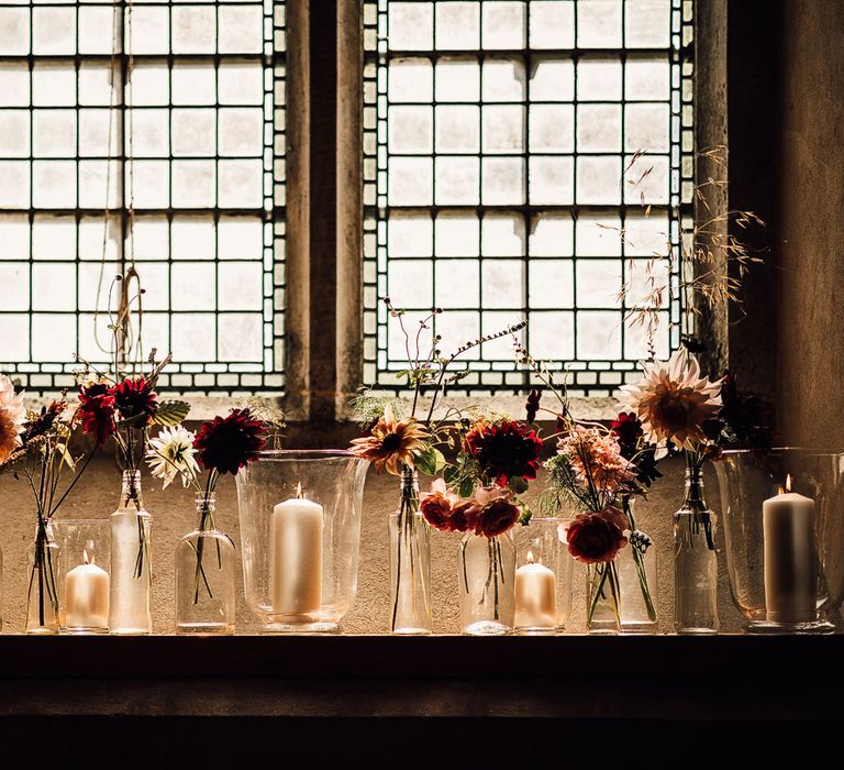 Mixed candles in glass candle holders and pink and red florals in assorted glass vases on church windowsill for Cornish wedding