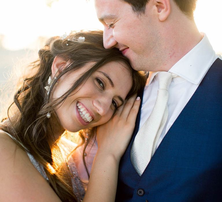 Bride leans into grooms chest on her wedding day as the sun shines around them and they both smile lovingly