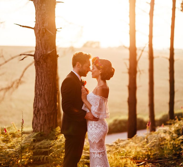 Bride in off the shoulder Grace Loves Lace wedding Dres sand flower crown stands with groom in black suit in woodland during golden hour