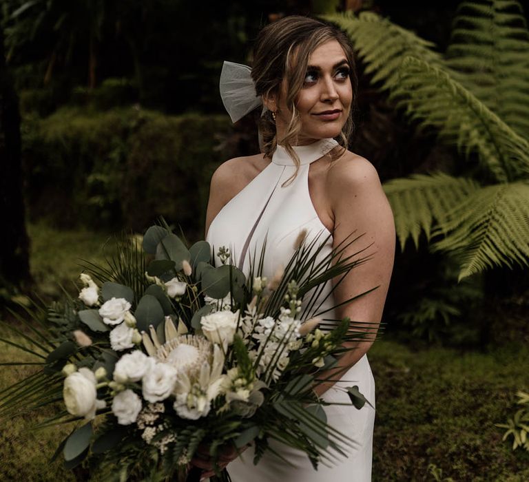 Bride in a fitted halterneck wedding dress holding a white flower and green foliage wedding bouquet 