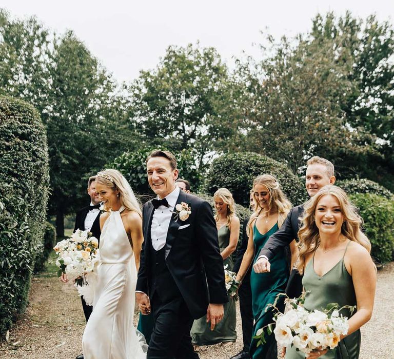 Bride in halterneck Halfpenny London wedding dress and veil holding hands with groom in black tie walks with bridesmaids in green satin bridesmaid dresses