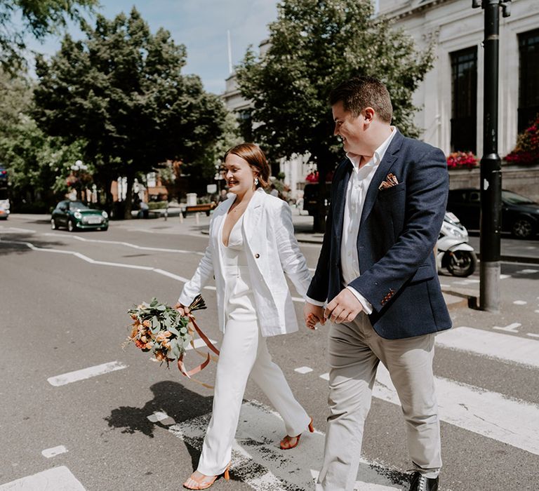 Bride in a jumpsuit and jacket holding hands with her groom in a blue blazer as they cross the road together 