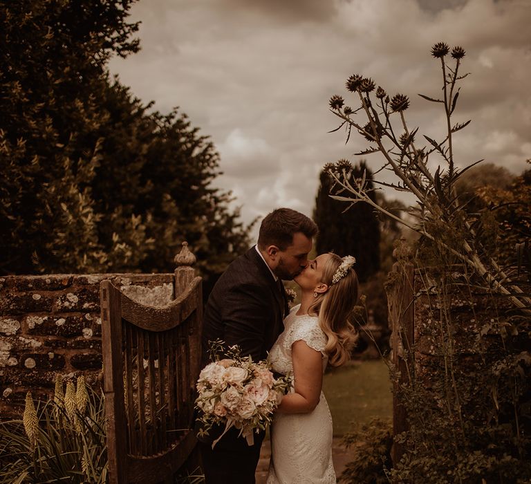 Bride in a lace Jesus Piero wedding dress and flower crown kissing her groom in the gardens at Dewsell Court, Hereford wedding venue  