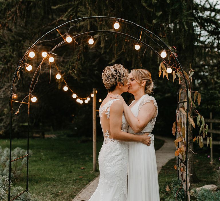 Brides kiss outdoors under festoon lighting on their wedding day