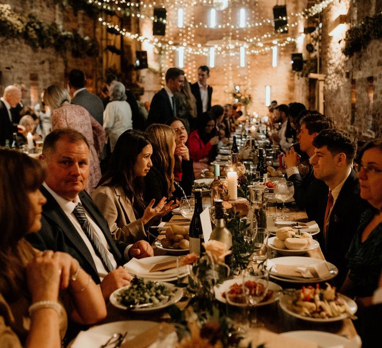 Wedding guests have dinner at rustic wedding breakfast at long wooden tables in stone barn with fairy lights, lit candles and rustic wedding table decor