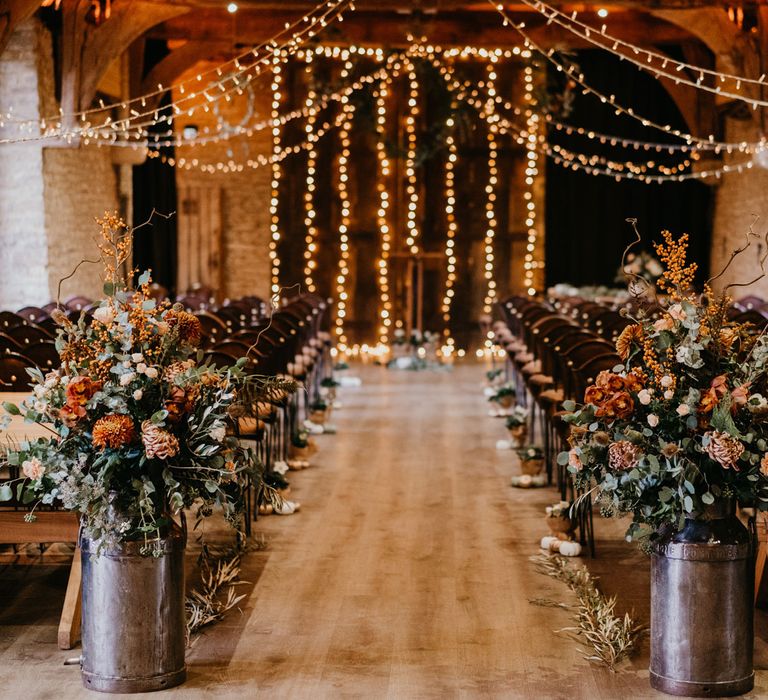 Rustic wedding aisle with milk churn floral installations, fairy light canopy and wooden chairs for rustic wedding ceremony at The Tythe Barn in Launton