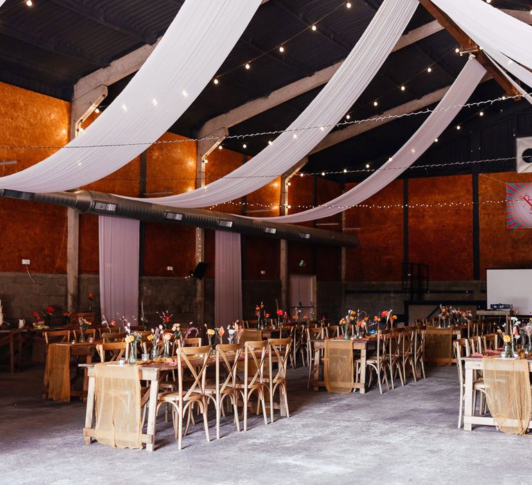 Barn festival style wedding reception with long wooden tables, orange florals, white draping and fairy lights