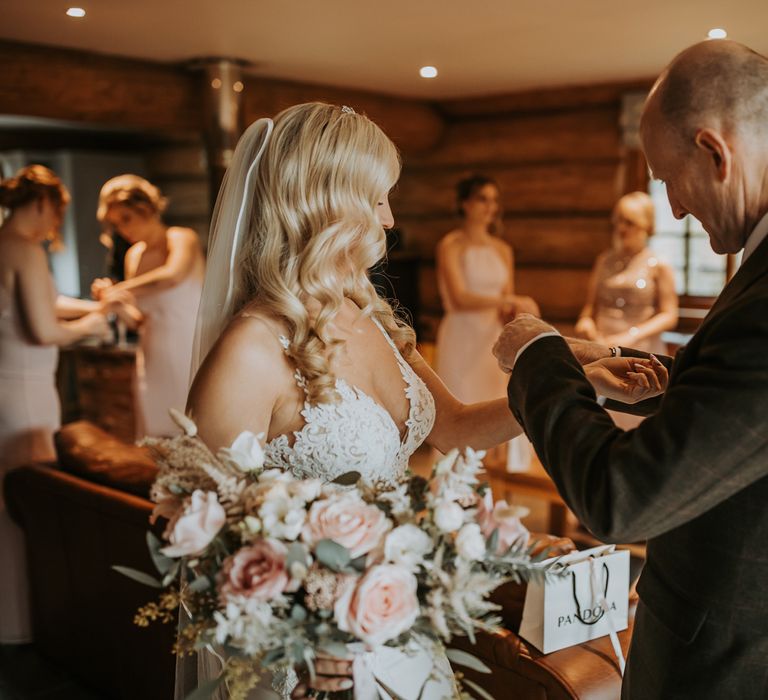 Bride with curled blonde hair, lace wedding dress and veil holding pink and white bridal bouquet has bracelet put on by father of the bride