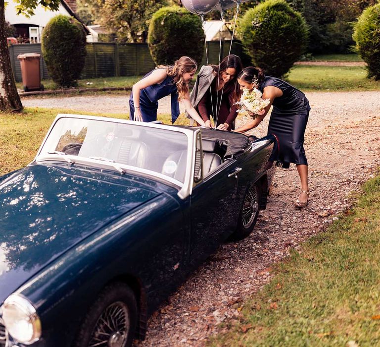 Bridesmaids in navy satin bridesmaid dresses attach balloons to dark blue vintage convertible