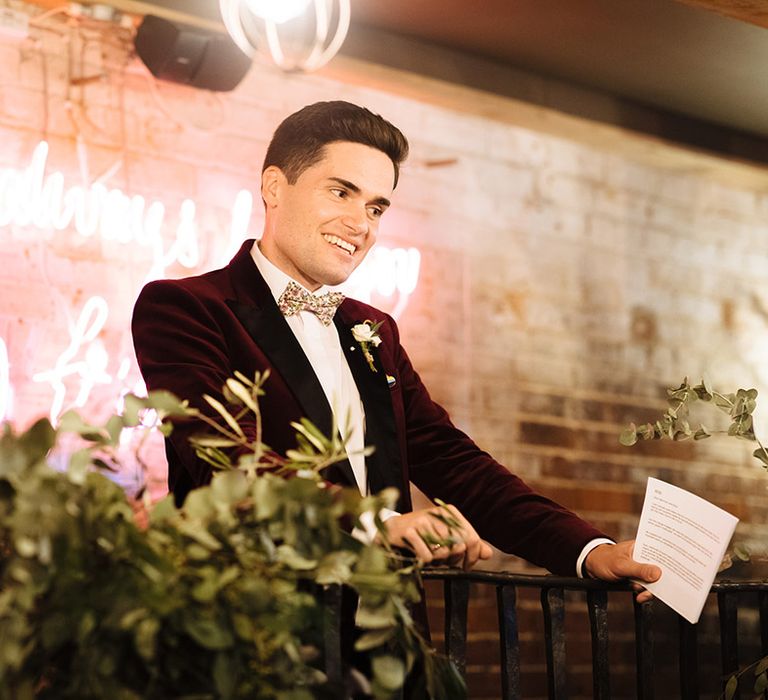 Groom in burgundy suit jacket reads out his wedding speech in front of pink neon sign at venue in Essex