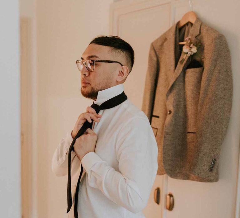 Groom tying black suit tie wearing crisp white long-sleeved shirt 