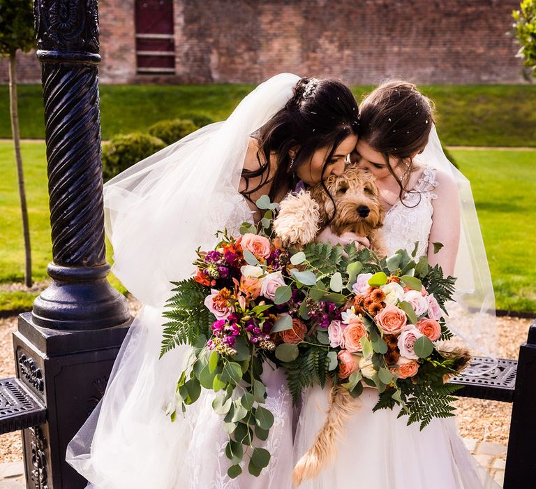 Brides in white wedding dresses holding matching wedding bouquets kiss their pet dog who was ring bearer during their ceremony