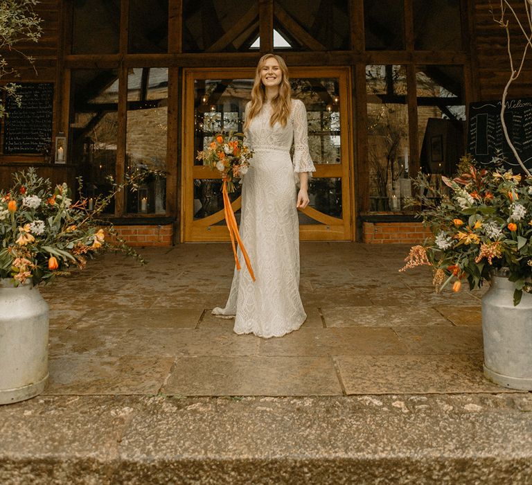 Bride in full lace a-line Rosa Clara wedding dress with bell sleeves and orange autumnal bouquet in front of Nancarrow Farm 