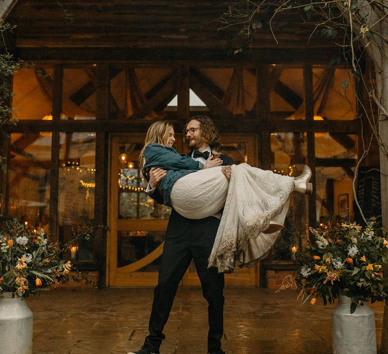 Groom in black tie lifts up bride in lace wedding dress with embroidered denim jacket