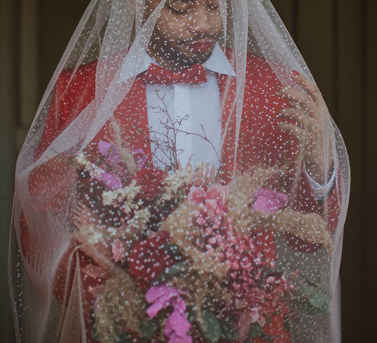 Groom in a red suit and bow tie wearing winged eyeliner and an embroidered veil over his face 