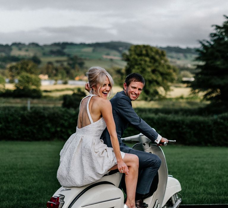 Bride in short wedding dress and groom in dark grey suit on white wedding vespa 